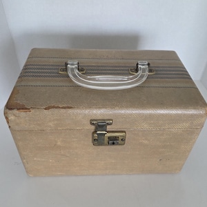 May include: Tan vintage suitcase with a clear plastic handle and a metal clasp. The top of the suitcase has three dark blue stripes. The suitcase is rectangular and has a textured surface. The suitcase is closed.