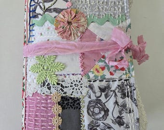 May include: A handmade journal with a patchwork cover. The cover features various fabric patterns, including floral prints, lace, and textured fabrics in shades of pink, gray, and white. A pink ribbon wraps around the journal, and a beaded charm hangs from the bottom.