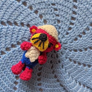 以下が含まれることがあります： 帽子をかぶったクマの形をした、かぎ針編みで作ったぬいぐるみです。クマは赤色で、顔は黄色と黒です。帽子はベージュで、赤い縁取りがあります。クマは青い編み物の毛布の上に横たわっています。