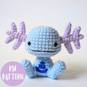May include: A handmade crochet plush toy in light blue with lavender appendages. The toy has black eyes and a smiling mouth. A blue design is on the chest. The image includes a purple circle with the text "PDF PATTERN".