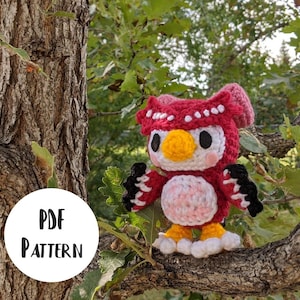 Puede incluir: Un juguete de peluche de búho tejido a crochet con detalles rojos, blancos y negros. El búho tiene una cara rosa y un pico amarillo. El juguete está sentado en una rama de árbol.