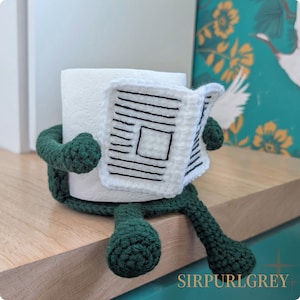 Puede incluir: Un original portarrollos de papel higiénico hecho a mano. El soporte es un rollo de papel higiénico blanco abrazado por un cuerpo de ganchillo verde con brazos y piernas. Parece estar leyendo un periódico de ganchillo con líneas negras y un cuadrado.