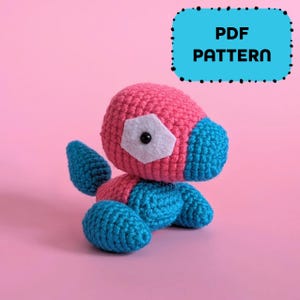 Pode incluir: Um brinquedo de crochê feito à mão em rosa e azul, com um detalhe em feltro branco e um olho preto. O brinquedo está sentado. Um retângulo azul com o texto "PDF PATTERN" está no fundo.