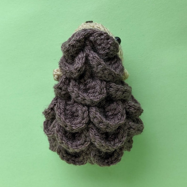 May include: A brown crocheted hedgehog with a textured, scaly back. The hedgehog is made with a soft yarn and has a cute, whimsical appearance.
