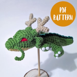 Puede incluir: Dragón de crochet verde con ojos rojos y garras blancas. El dragón tiene cuernos y una cola enroscada. El dragón está en un palo de madera y una base de madera. La imagen tiene el texto "PDF PATTERN" en un círculo amarillo.