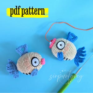 Puede incluir: Dos juguetes de pez tejidos a ganchillo con cuerpos beige, aletas y colas azules, y labios rosas. Un pez tiene un gorro rojo y un hilo naranja. El texto "pdf pattern" está arriba y "sirpurlgrey" está escrito en blanco en la parte inferior.