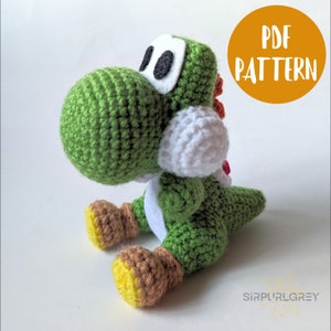 Könnte beinhalten: Grünes gehäkeltes Yoshi Plüschtier mit weißen Augen, einem weißen gehäkelten Lätzchen und braunen gehäkelten Füßen. Das Spielzeug sitzt auf einer weißen Oberfläche. Der Text "PDF PATTERN" befindet sich in einem Kreis auf der rechten Seite des Bildes.