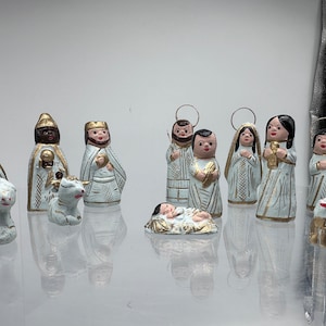 Könnte beinhalten: Eine Weihnachtskrippe mit 12 Figuren, darunter Maria, Josef, Jesuskind, drei Könige und Hirten. Die Figuren sind aus weißem Ton mit goldenen Akzenten und etwa 7,5 cm hoch.