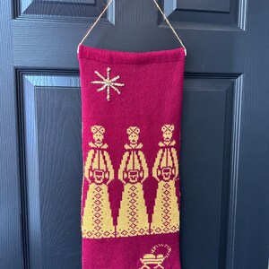 May include: A red knitted wall hanging with a gold star and three figures in gold singing carols. The bottom of the hanging has a gold manger with a star.