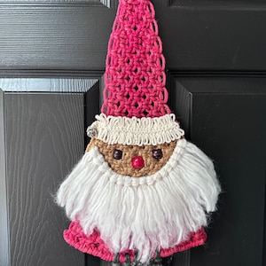 May include: A crocheted gnome ornament with a pink hat, white beard, and brown face. The gnome is hanging on a black door.