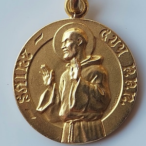 Puede incluir: Pendiente de color dorado con una representación de un santo, posiblemente San Francisco, con la inscripción "Sanctus Franciscus" alrededor del borde.