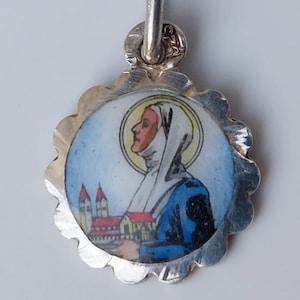 May include: Silver pendant with a colorful enamel image of a woman in a blue robe and white head covering. The image is surrounded by a scalloped silver border.