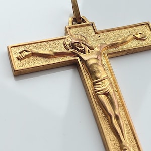 Puede incluir: Colgante de crucifijo dorado con una detallada representación de Jesucristo. La cruz tiene un borde texturizado y un lazo para colgar. La figura de Cristo está con un taparrabos drapeado.