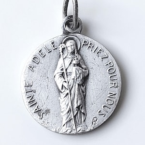 Puede incluir: Un colgante redondo de metal plateado con una imagen en relieve de una figura que sostiene un bastón. El texto "SAINTE ADELE PRIEZ POUR NOUS" está grabado a lo largo del borde circular. El colgante incluye una pequeña anilla metálica en la parte superior.