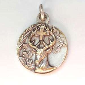 May include: A circular silver-toned metal pendant featuring a raised relief of a deer head with large antlers. A small gold-toned cross is positioned between the antlers. The design includes detailed oak leaves on the sides and a metal bail at the top.
