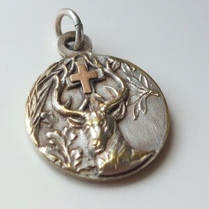 May include: A circular silver-toned metal pendant featuring a raised relief of a stag head with antlers positioned beneath a small copper-colored cross. The design includes detailed foliage elements on the sides and a silver jump ring at the top.