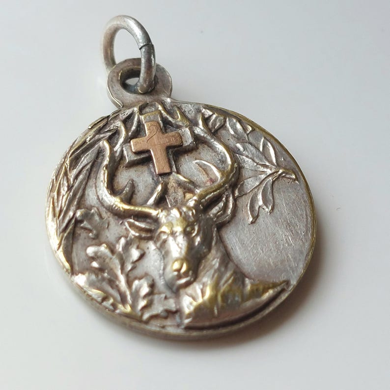 May include: A circular silver-toned metal pendant featuring a raised relief of a stag head with antlers positioned beneath a small copper-colored cross. The design includes detailed foliage elements on the sides and a silver jump ring at the top.