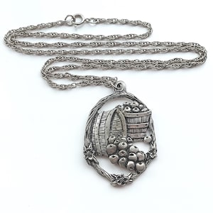 May include: Silver-toned pendant necklace featuring a detailed design of a barrel and a basket overflowing with fruit. The pendant is surrounded by a floral frame and hangs from a delicate chain. The necklace is a vintage style.