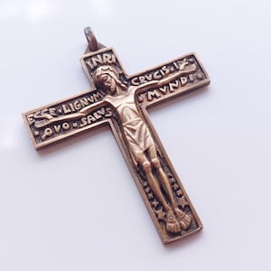 May include: A bronze-toned metal crucifix pendant featuring a central figure of a person on a cross. The top of the cross displays the letters INRI, while the horizontal arms are inscribed with the Latin text ECCE LIGNVM CRVCIS and SALVS MVNDI.