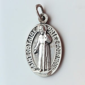 Może przedstawiać: Owalny srebrny wisiorek przedstawiający postać religijną w szacie, trzymającą laskę. Wisiorek ma napis "SAINT ORTAIRE PRIEZ POUR NOUS". Posiada pętlę u góry do przymocowania do łańcuszka.