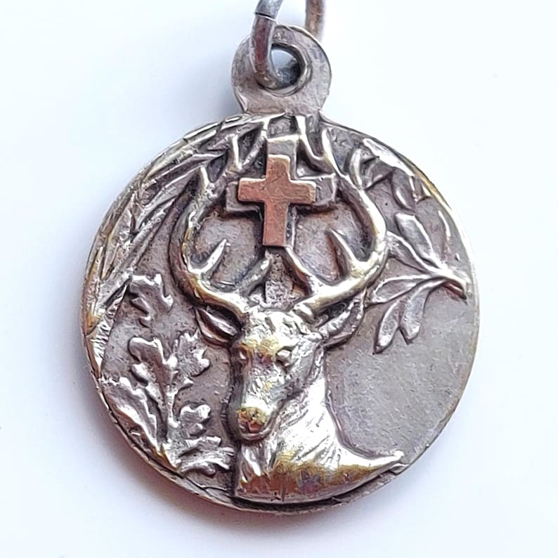 May include: A circular silver-toned metal pendant featuring a raised relief of a stag head with antlers. A small cross is positioned between the antlers. The design includes oak leaves on the left and right sides. A metal loop is attached at the top.