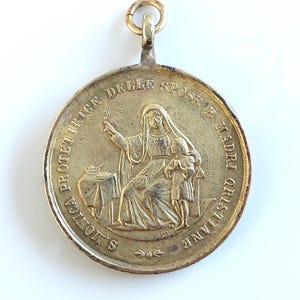 May include: A round gold-toned metal religious pendant featuring a raised relief of Saint Monica with a child. The inscription reads S. MONICA PROTETTRICE DELLE SPOSE E MADRI CRISTIANE. The pendant includes a small circular bail at the top for hanging.