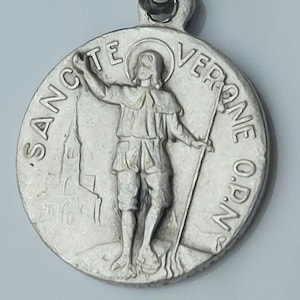 Op de afbeelding: Zilverkleurig metalen hanger met een religieus figuur met een staf, omringd door de inscriptie "SANCTE VERONE O.P.N."