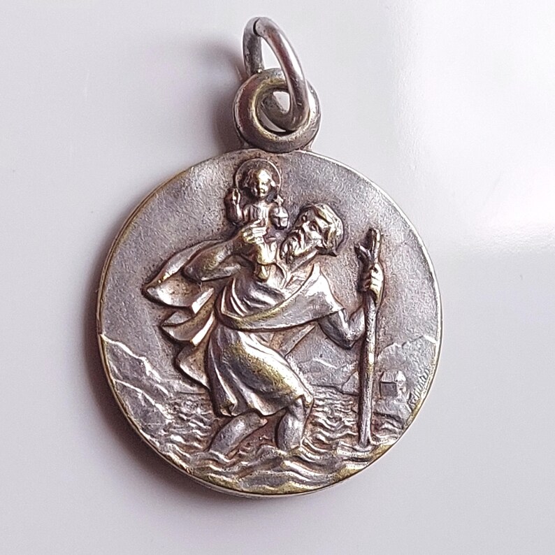 May include: A round silver-toned metal pendant featuring a relief depiction of Saint Christopher carrying the Christ Child on his shoulder while walking through water with a staff. The pendant includes a circular metal jump ring at the top.