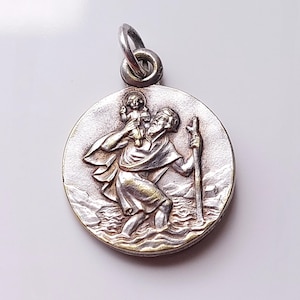 May include: A round silver-toned metal pendant featuring a relief design of a bearded figure carrying a small child on their shoulder while walking through water with a staff. The pendant includes a circular jump ring at the top for attachment.