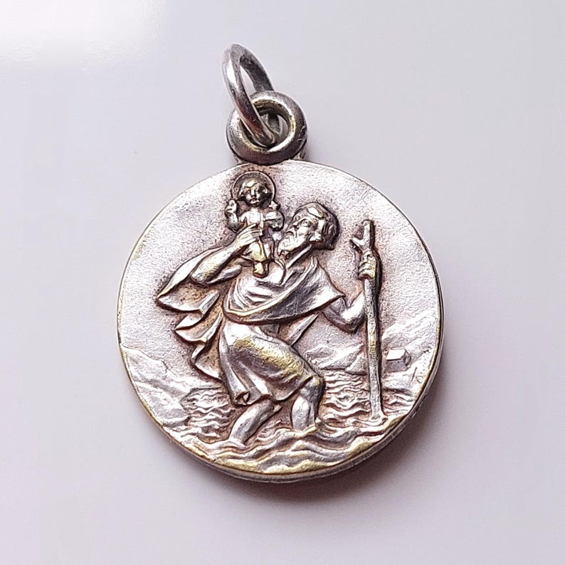 May include: A round silver-toned metal pendant featuring a relief design of a bearded figure carrying a small child on their shoulder while walking through water with a staff. The pendant includes a circular jump ring at the top for attachment.