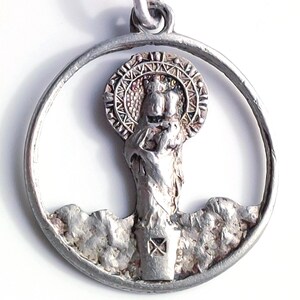 May include: A silver pendant featuring a religious figure within a circular frame. The figure, possibly a saint or deity, is depicted with a halo and stands atop a textured base. The pendant is likely made of a metal alloy.