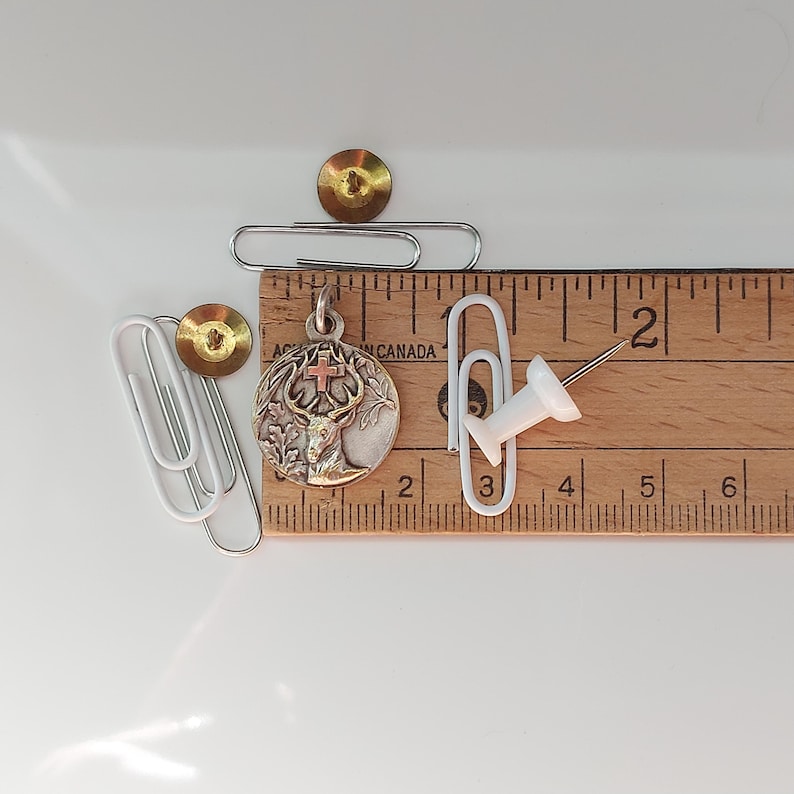 May include: A circular silver-toned metal pendant featuring a relief of a stag with a cross between its antlers, positioned next to a wooden ruler showing a diameter of approximately 1 inch, alongside white paper clips and a white push pin.