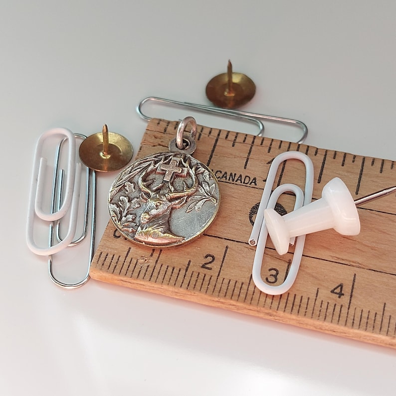 May include: A circular silver-toned metal pendant featuring a relief of a stag with a cross between its antlers, surrounded by oak leaves. The pendant is placed on a wooden ruler marked in inches, alongside two brass thumbtacks, a white thumbtack, and three silver-toned paper clips.