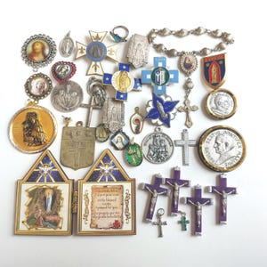 May include: A selection of religious pendants, crosses, and medallions. The items display religious figures, crosses, and text. The colours include gold, silver, blue, and purple. Various shapes and sizes are present.