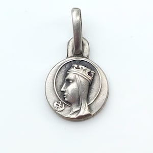 May include: A circular silver-toned metal pendant featuring a relief profile of a figure wearing a crown. The design includes a small hexagonal emblem near the chin of the figure and a sturdy vertical loop at the top for attachment to a chain.