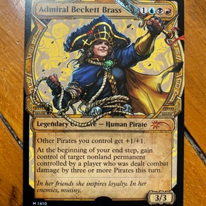 Puede incluir: Admiral Beckett Brass, una carta de Magic: The Gathering que representa a una pirata humana legendaria con un atuendo azul y dorado, sosteniendo una espada y una cuerda. El texto de la carta dice "Los demás piratas que controlas obtienen +1/+1. Al comienzo de tu paso final, gana el control de un permanente no tierra objetivo controlado por un jugador que haya recibido daño de combate de tres o más piratas este turno. En sus amigos inspira lealtad. En sus enemigos, motín."