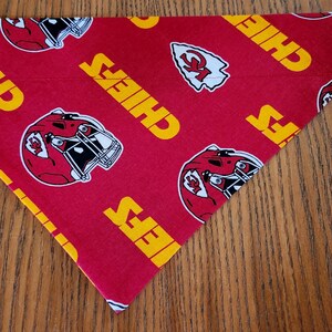 May include: Red fabric dog bandana with a white and yellow pattern of the Kansas City Chiefs logo and the word "Chiefs" repeated. The back of the bandana is plain red fabric.