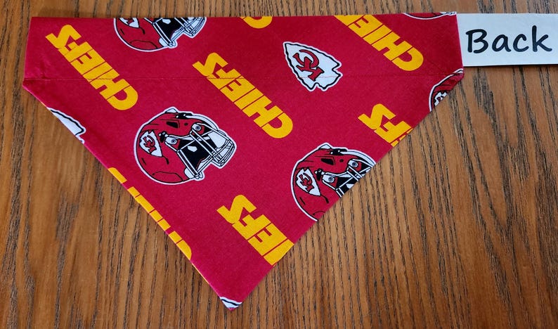 May include: Red fabric dog bandana with a white and yellow pattern of the Kansas City Chiefs logo and the word "Chiefs" repeated. The back of the bandana is plain red fabric.