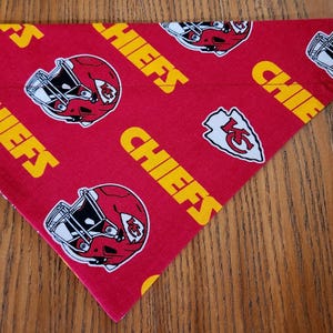 May include: Red, white, and yellow dog bandana with a repeated pattern of a football helmet and the word "Chiefs". The bandana is folded in a triangle shape and has the word "Front" written on a white tag.