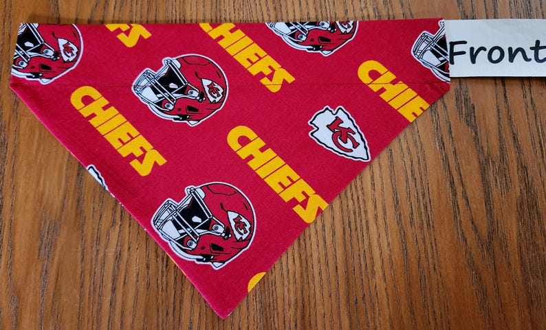 May include: Red, white, and yellow dog bandana with a repeated pattern of a football helmet and the word "Chiefs". The bandana is folded in a triangle shape and has the word "Front" written on a white tag.