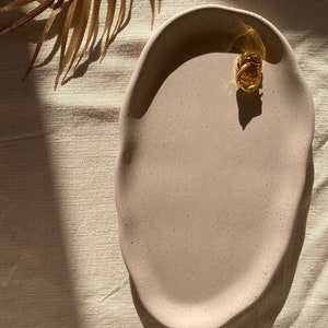 May include: A light pink oval ceramic serving tray with a speckled finish. The tray is on a beige linen tablecloth.