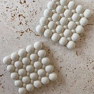 May include: Two white ceramic coasters with a textured bubble design. Each coaster features a pattern of raised circles.