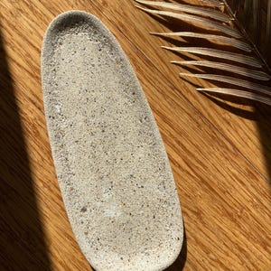 May include: A light beige, oval-shaped tray with a textured surface. The tray is made of a porous material and sits on a wooden surface. A dried palm leaf is in the background.