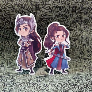 Pode incluir: Dois autocolantes recortados com personagens em estilo anime. Um usa armadura e capacete, o outro segura uma espada e usa uma roupa azul e vermelha. Ambos têm cabelo castanho comprido, sobre um fundo padronizado.