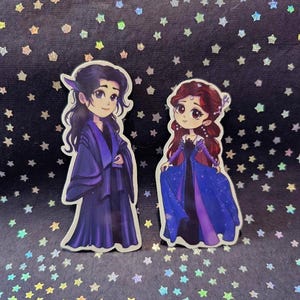 Peut inclure: Deux autocollants de style dessin animé représentant des personnages fantastiques. L'un porte une robe violet foncé, l'autre une robe bleue et violette. Le fond est une surface sombre parsemée de confettis en forme d'étoiles.