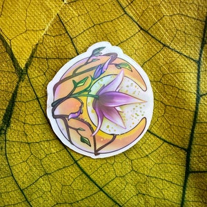 Puede incluir: Una pegatina con una luna creciente con una flor morada que crece de ella. La luna está rodeada de un brillo dorado y la flor tiene un centro blanco. La pegatina está sobre un fondo de hoja amarilla.