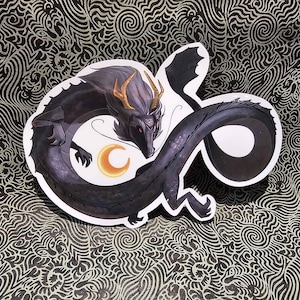 Puede incluir: Una pegatina en blanco y negro de un dragón enroscado alrededor de una luna creciente. El dragón tiene un cuerno dorado y está sobre un fondo de remolinos.