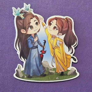 May include: A sticker featuring two anime-style characters. One character wears a blue dress and has a crown. The other character wears a yellow dress and is reaching up. A red and black instrument is held by the character in blue.