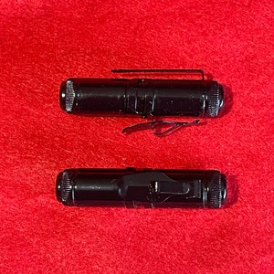 Puede incluir: Dos linternas de metal negro con un clip en el lateral. Las linternas están sobre una superficie roja.