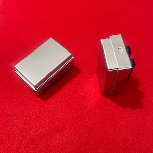 May include: Two small, rectangular, silver metal boxes on a red surface. One box is closed, and the other is open. The open box is slightly angled, revealing its interior. The second box has a black component attached to it.